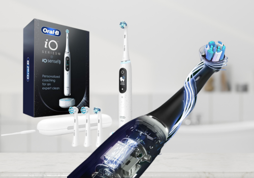 The image displays the Oral-B iO Series electric toothbrush with its packaging and accessories. The blue retail box highlights "DEEP CLEAN" and "400% more PLAQUE REMOVAL". A light blue toothbrush is shown on its charging base, along with a white travel case and an extra brush head. A promotional close-up illustrates the toothbrush removing plaque, claiming "Remove 100% more plaque in 1 week*". The Oral-B logo is visible. The setting is a softly blurred bathroom.